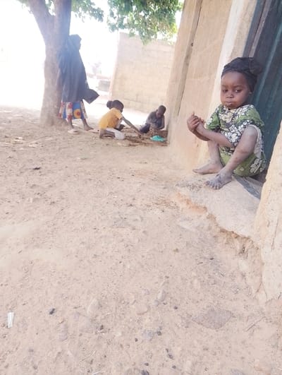 This image captures an outdoor scene in Kanwa, Nigeria. In the immediate foreground, a young child, approximately 2-4 years old, sits on a raised concrete ledge of a light-colored, possibly mud-brick or block, building on the right. The child is wearing a green and white patterned dress, a dark headwrap, and has visible dirt on their bare feet and lower legs. Their hands are clasped in front, and they are looking directly forward with a neutral expression.
In the middle ground, to the left of the scene, three other individuals are present on a dry, light brown dirt ground containing small rocks and sparse debris. To the far left, partially obscured by the trunk of a tree, stands an individual wearing a dark, long-sleeved outer garment over patterned clothing, with white sandals visible on their feet. Closer to the center, two other children are crouched or sitting on the ground. One child, wearing a yellow shirt and light-colored shorts, is actively manipulating or playing with dirt and small objects on the ground with their hands. Adjacent to this child, a second child, dressed in darker attire, is also focused on the ground, near a light blue object resembling a small bowl or container. The background features more structures made of light-colored blocks or packed earth. The scene is illuminated by natural daylight.