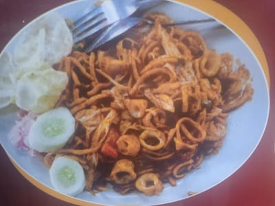 A close-up, overhead shot shows a plate of mie goreng, or fried noodles, garnished with sliced cucumbers and a side of what appears to be crackers and pickled vegetables. The noodles are dark brown and coiled, interspersed with visible rings of squid. A fork is partially visible, resting on the noodles. The plate is white and sits on a dark red surface with a yellow border. The image depicts a food item that might be found at a street food stall or restaurant in Peurupok, Indonesia, a coastal village known for its fresh seafood, making the inclusion of squid a likely culinary element.