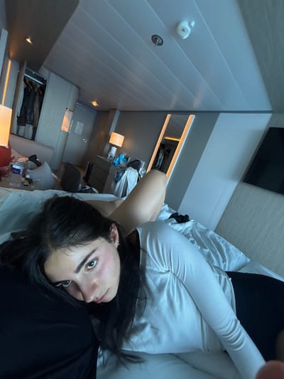The image depicts an indoor scene within a modern cabin or hotel room, viewed from a low angle. The specific location within San Miguel de Cozumel, Mexico, cannot be determined from visual evidence in the image.
In the foreground, a young female individual is partially reclining on a bed with white bedding, her head resting on a dark pillow. She has long, dark hair and dark eyebrows, and her gaze is directed towards the camera. She is wearing a light-colored, long-sleeved top and dark lower garments. Partially obscured behind her on the bed, a bare leg is visible.
The room features a white, paneled ceiling with recessed lighting fixtures and a white smoke detector/sprinkler unit. To the left, an open wardrobe reveals various dark garments hanging inside. A bedside table next to the bed holds a lamp, a red spherical object, a clear water bottle, and other small containers. In the mid-background, a dresser with multiple drawers supports another lamp with a white shade, along with scattered items including bags and articles of clothing. Adjacent to the dresser, a large, vertically oriented mirror with illuminated edges reflects an additional area of hanging clothes. A dark, rectangular screen, consistent with a television, is mounted on the wall to the far right. A closed, light-colored door is visible in the central background. The room appears to be in a state of casual occupancy.