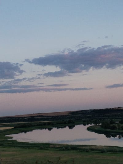 This media file depicts a tranquil landscape scene at dusk in Bilousivka, Ukraine. The sky transitions from a pale blue at the top to soft pink and lavender hues closer to the horizon, suggesting the late evening hours. Scattered clouds, tinged with purple and pink, drift across the sky.
The foreground features a winding river with calm water reflecting the sky and surrounding greenery. The riverbanks are lined with lush green grass and dark trees, creating a stark contrast with the muted tones of the sky. Beyond the river, rolling hills rise, covered in a mix of fields and darker vegetation. The overall impression is one of peace and quiet.
There is no discernible activity or event taking place; the image captures a static, natural setting. The time of day is clearly indicated by the twilight sky. The weather appears to be clear, with the soft light suggesting a mild evening. No people or objects are visible in the frame, emphasizing the natural beauty of the location. There is no visible text in the image.