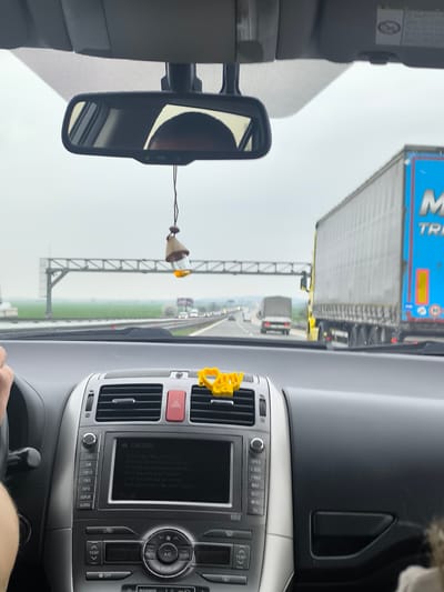 The image depicts the interior of a moving vehicle, observed from the driver's perspective, looking forward through the windshield onto a multi-lane highway near Opalchenets, Bulgaria.
Inside the vehicle, the lower portion of a grey and black dashboard is visible. A central infotainment system displays a "CAUTION" message on its screen, flanked by silver control buttons for audio and navigation functions, and climate controls featuring "AUTO," "A/C," and "DUAL" buttons. A red triangular hazard light button is present. A small yellow heart-shaped object is clipped to an air vent on the passenger side of the dashboard. The driver's left hand and forearm are visible on the left, positioned near the steering wheel. A centrally mounted rearview mirror reflects the upper portion of a person's head, presumably the driver's. A small, teardrop-shaped air freshener containing yellow liquid hangs from the rearview mirror.
Outside the vehicle, the highway features at least two lanes of traffic. A large yellow and blue semi-trailer truck, partially displaying "M TRI" on its side, is prominent on the right, appearing to move alongside or slightly ahead of the observer's vehicle. Further down the highway, another white truck and several smaller vehicles are visible. A large, grey metal gantry spans across the highway in the mid-distance. The surrounding landscape consists of expansive green fields extending to the horizon under an overcast sky. White concrete barriers line the highway's median and shoulders.