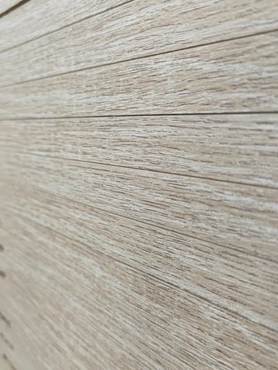 The image is a close-up, full-frame, eye-level shot of a textured surface resembling light-colored wood planks. The planks are horizontally oriented and run from the bottom left to the top right of the frame, creating a sense of depth due to the slight angle. The texture displays a prominent grain pattern with lighter and darker striations, suggesting a natural or laminate wood finish. There are no people, animals, or other objects visible in the image. The lighting appears even, highlighting the texture of the surface. The background is entirely composed of this wood-like material. The location is not identifiable from the image.