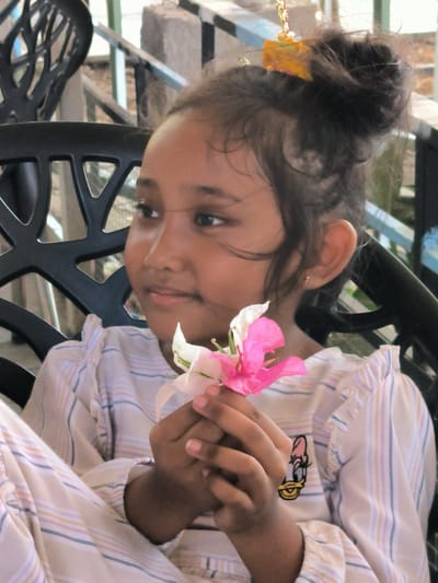 A young girl is seated, holding a pink and white flower. She is wearing a striped shirt with a cartoon character on it, and her hair is up in a bun. The background shows outdoor seating and a structure, indicating an outdoor or semi-outdoor setting. The location is Matang, Indonesia.