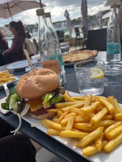 The image depicts an outdoor dining scene in Porto Cristo, Spain, focusing on food and beverages at a table with a waterfront background. In the immediate foreground, a plate holds a large burger and a generous serving of golden-brown french fries. The burger features a light brown bun with a patterned top, held by a wooden skewer, encasing a patty, melted yellow cheese, lettuce (both green and red-tinged), a red tomato slice, and white onion rings. To the left, a smaller plate contains additional french fries.
To the right of the main food plate, a clear glass filled with sparkling water, ice, and a lemon wedge is visible. Behind it, a small, circular white ashtray contains two cigarette butts. Two tall, clear glass bottles stand on the table; one has a blue label visible at its base, and the other, slightly behind it, has an orange-brown label. Further back on the table, a blurred pizza with a cheese topping is visible.
The table surface is dark. On the left side of the frame, a person wearing a dark maroon or purple top is seated, partially visible and blurred, facing away from the camera. The lower left corner shows a partial view of a person's dark-clad lap.
The background reveals a bustling outdoor restaurant environment. Several blurred tables and chairs are visible under large, light-colored parasols. Beyond the restaurant, a harbor or marina stretches, populated with boats and white buildings lining the waterfront. Hilly terrain rises in the distance under a partially cloudy sky. The overall scene suggests a bright, daytime setting.