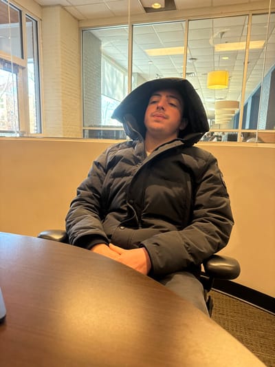 A person wearing a black puffer jacket with the hood up sits at a brown table. Light from a window illuminates the left side of the frame, where the window provides a view of trees and buildings. Overhead, modern pendant lights are visible. The person is seated on a black chair.