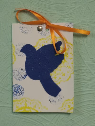A handmade gift tag, likely created for a child, is displayed on a textured light green surface. The tag is rectangular and oriented vertically. The front of the tag features a large, dark blue silhouette of a bird, positioned slightly off-center to the left. The bird appears to be cut out and adhered to the tag, with a subtle glitter effect visible on its surface. The background of the tag is white and decorated with a pattern of yellow and blue stamps. The yellow stamps are circular and appear to be stylized flower-like shapes, while the blue stamps are smaller and more abstract, resembling clusters of small dots or scribbles. A silver brad is fastened through the top of the tag, securing an orange ribbon tied in a bow. The ribbon forms a loop, suggesting the tag is intended to be hung. The overall aesthetic is whimsical and crafted with simple materials. The image was taken in Khotsky, Ukraine.