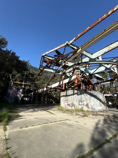 The image shows a dilapidated, rusty metal structure, possibly part of an abandoned cable car system, set against a clear blue sky and a backdrop of green trees. The structure's steel frame is heavily corroded with significant rust, suggesting long-term disuse and exposure to the elements. The structure is located in San Antonio de Galipán, Venezuela. It is situated next to a concrete pathway or platform. The image is rotated counter-clockwise. A concrete wall adorned with graffiti is visible.