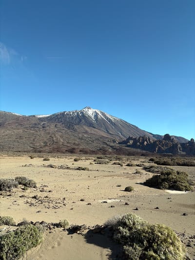 The image presents a scene in Paradores Cañadas del Teide, Spain. A large mountain with some snow at its peak dominates the left side of the frame, contrasting against a clear blue sky. To the right of the mountain, a dry, sandy terrain is visible, sparsely populated with low-lying vegetation. The landscape appears arid and rocky.