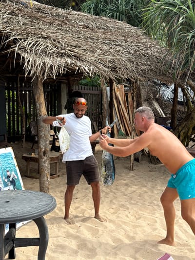 Two men display their catches. One, shirtless in blue shorts, holds a long fish, a knife in his other hand. The other, wearing a white t-shirt, brown shorts, and sunglasses, shows off a smaller fish. They are on a sandy beach near a wooden structure with a thatched roof. A table and chair are partially visible in the foreground. The setting appears tropical.