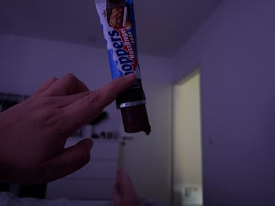 A person's hand is holding a half-eaten chocolate bar called "Knoppers Nussriegel Dark." The chocolate bar has a blue and white wrapper with the brand name prominently displayed. The person's thumb is extended, pointing towards the chocolate bar. The background is a dimly lit room with a purple wall and a partially visible white dresser. There is a doorway in the background leading to a lighter area. The image appears to have been taken indoors, likely in a residential setting within Kirtorf, Germany, given the context of the request.