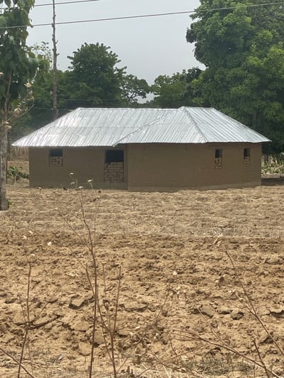 The image, oriented incorrectly on its side, depicts a rural landscape under bright daylight. A simple, single-story building with a reflective, corrugated metal roof is prominent on the left side of the frame. Its walls appear to be constructed from earthen materials, possibly mud bricks, and are a light brown or tan color. Several small, rectangular openings, likely windows, are visible in the building's walls; some of these openings are partially blocked with stacked bricks or stones.
To the right of the building, a large expanse of dry, cracked, and uneven earth dominates the midground and foreground. This barren ground suggests a dry season or an uncultivated field. Scattered across this dry earth are thin, dry branches and plant stalks, indicating sparse vegetation.
In the background, beyond the building and the dry field, a dense canopy of lush, green trees extends towards a bright, hazy sky, which occupies the upper left portion of the image. The overall impression is one of a rustic dwelling situated in a dry, possibly arid, environment with more verdant areas in the distance.
No people or animals are visible, and no specific activity is taking place. The scene conveys a sense of stillness and the natural conditions of the area. The clear sky and strong lighting suggest it is daytime, likely mid-morning or early afternoon. There is no visible text in the image. The architectural style and arid landscape cues align with the provided location of Unguwar Gaya, Nigeria.
