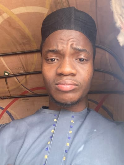 The image presents a close-up portrait of an adult male of African descent, positioned centrally within the frame. He wears a black, knitted kufi cap and a grey traditional tunic with a band collar, featuring vertical embroidery composed of small, square-like yellow and blue/purple elements on either side of the chest placket. His expression is neutral to slightly pensive, with mildly furrowed eyebrows and gently pursed lips. He has faint facial hair visible as stubble on his chin and jawline and looks directly at the camera. The background reveals the interior ceiling of a vehicle, possibly a commercial tricycle (keke napep), characterized by light-colored paneling or fabric adorned with a pattern of intersecting curved lines in shades of brown, yellow, red, and blue. Two dark, horizontal metal support bars are visible across the upper portion of the background. The scene is located in the city of 0801712591, Nigeria.