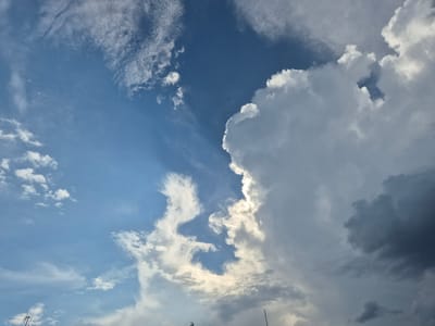 This is a close-up, low-angle shot of a dynamic sky above Rzeszów, Poland. The sky is a vibrant blue with a mix of cloud formations. On the right, a large, dense cumulus cloud dominates the frame, its edges brightly illuminated by sunlight, casting deep shadows on its underside. To the left of this massive cloud, the sky transitions to a clearer blue, scattered with thinner, wispy cirrus clouds that stretch across the expanse like brushstrokes. Towards the lower edge of the image, faint silhouettes of what appear to be antenna or utility poles are visible against the bright clouds, suggesting an urban or semi-urban environment. The overall impression is one of a dramatic and changing weather system.