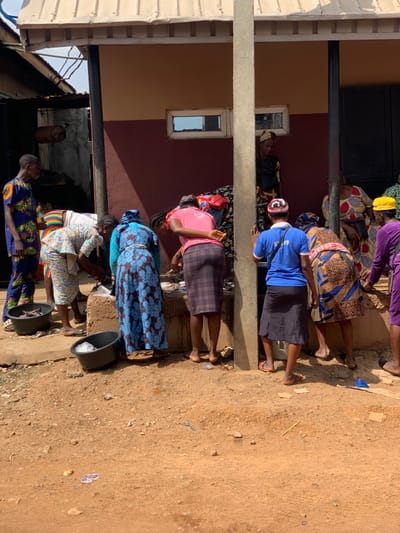 A group of women and girls are standing in front of a building in Ondo, Nigeria. The building is a single story structure with a brown brick facade. It has a large, covered porch that extends along the front of the building. Several women are gathered outside the building, likely waiting for something. The women are dressed in brightly colored traditional Nigerian clothing and are interacting with each other. There is a large black bucket on the ground next to them. There is also a lot of loose dirt and debris on the ground in front of the building. The ground is mostly dry and dusty. The scene is located in a residential area in Ondo, Nigeria.
