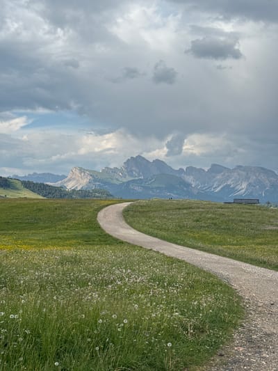 This media file depicts a scenic mountain landscape with a walking path. The subjects include rugged, imposing mountains in the background, a lush green meadow dotted with small white flowers in the foreground, and a winding gravel path that curves through the meadow. A small, dark structure, possibly a bench or shed, is visible on the slope of the mountain in the middle ground. The sky is filled with dramatic, cloudy formations, suggesting a dynamic weather system. The overall impression is one of natural beauty and tranquility, with the path inviting exploration of the vast mountain scenery. The time of day appears to be daytime, likely late morning or early afternoon, given the lighting conditions. The location, Kastelruth - Castelrotto, Italy, is known for its stunning alpine vistas. No text is visible in the image.