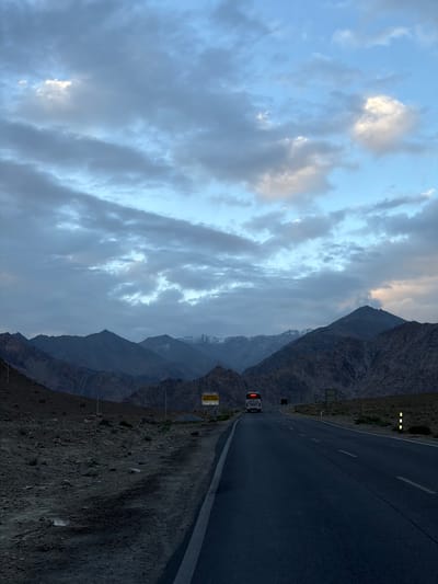 A van drives on a paved road winding through a mountainous landscape. The sky is filled with dramatic clouds, with patches of sunlight breaking through, suggesting either sunrise or sunset. The mountains are rugged and appear to be barren, with some snow visible on their peaks. The road is dark asphalt with white lane markings. The van's brake lights are illuminated. There are some lights visible along the side of the road, possibly streetlights or markers. A yellow sign with text in an unknown language is mounted on a pole next to the road. The scene evokes a sense of vastness and solitude, characteristic of a remote mountain region. The time of day appears to be either early morning or late evening, given the lighting conditions. The weather seems to be partly cloudy. The location is identified as Chumkar, India.