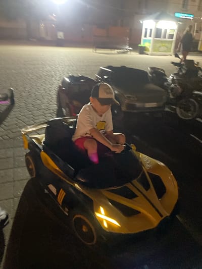 The image shows a child wearing a hat and a t-shirt, sitting in a yellow and black toy car. The child is holding onto a steering wheel. In the background, there are other toy cars, a motorcycle, a small building with illuminated windows, and a paved area. The lighting suggests it is nighttime or evening. The scene appears to be an outdoor recreational area in the city of Merchanskoe, Russia.