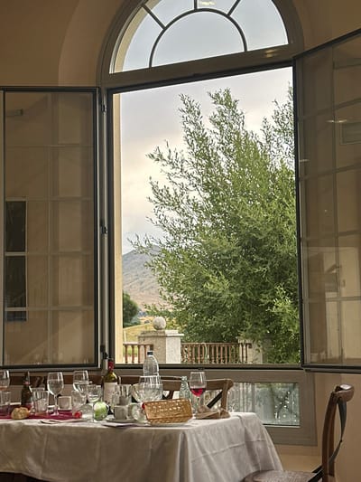 An open window frames a landscape view with a large tree and a distant mountain range under a cloudy sky. The window is adjacent to a semi-circular window. Part of a dining table, draped with a white cloth and laden with glassware and a bottle of wine, is visible in the foreground. The scene appears to be indoors, with the focus drawn to the outdoor scenery.