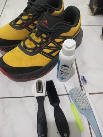 A pair of Eiger shoes, yellow with black trim and red soles, sits on a tiled surface. Beside the shoes are cleaning supplies: a bottle of "holy shine Shoe Cleaner", two black-handled brushes, a light blue toothbrush, and a green-handled pointed tool. The items appear arranged for shoe cleaning.