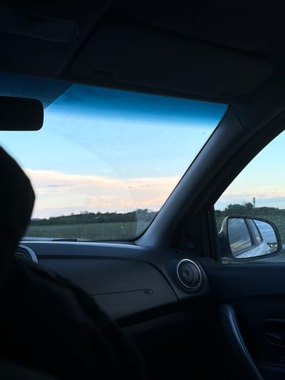 The image shows the interior of a car, looking out through the windshield. A person's dark silhouette is visible in the foreground on the left. The sky is a light blue with some wispy white and pink clouds, indicating either sunrise or sunset. A line of dark green trees forms the horizon in the distance, with a field in front of them. The car's dashboard is visible, including an air vent. A side mirror on the right side of the car shows a reflection of the road behind. The location is identified as Oktiabrskaya, Russia.