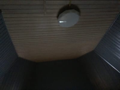 The image shows an indoor ceiling with wooden planks. A light fixture, possibly a ceiling lamp or a speaker, is suspended from the ceiling. The fixture is a light gray, multi-faceted shape. The overall lighting in the image is dim, with the exception of a bright reflection on the surface of the fixture. The location appears to be a residential interior, likely a home or a communal space within the SNT (SNT is a type of Russian horticultural non-profit partnership). No people or specific actions are depicted.