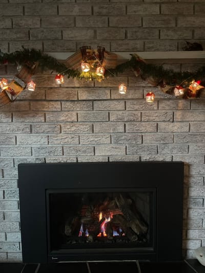 A decorated brick wall features a black-framed fireplace on the right side, where simulated flames flicker around faux logs. A Christmas garland adorned with ribbon bows and square-shaped lights descends vertically on the left side of the wall. The garland's soft glow adds warmth to the scene.