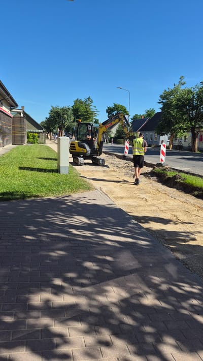 In the city of Līvāni, Latvia, on a bright, sunny day, a construction scene is depicted. In the foreground, a paved brick walkway with dappled shade from overhead trees leads the eye towards a construction area. A small yellow excavator is actively working on a road or sidewalk renovation project. A person wearing a high-visibility yellow vest is standing near the excavator, seemingly observing the work. The surrounding environment includes a grassy verge, a utility box, trees, and residential buildings visible in the background. The sky is clear blue. The overall impression is one of ongoing municipal work in a semi-urban setting.