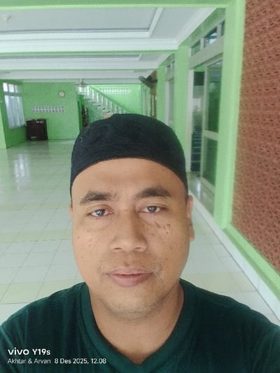 A selfie shows a man with dark hair, a dark skullcap, and a dark green shirt. He is inside a building with light green walls, a white ceiling, and tiled floors. Behind him, there is a staircase, a window, and an interesting wall design. The timestamp at the bottom left reads "vivo Y19s Akhtar & Arvan 8 Des 2025, 12.08," indicating the type of phone and date.