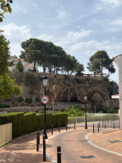 The image captures a scenic view of a street in Benalmádena, Spain, with a rocky cliff face prominently featured in the background. A winding brick-paved road, bordered by black bollards and a white line, curves through the foreground. A neatly trimmed green hedge runs along the left side of the road, separated by a low stone wall.

At the top of the cliff, several large pine trees and some palm trees are visible, along with white buildings with terracotta roofs. A dark opening, possibly a cave or tunnel, is present in the rock face. A street lamp with a decorative top stands near the road, accompanied by a circular speed limit sign displaying "30" within a red border. Another street lamp is visible further up the cliff. The sky is partly cloudy, with a mix of white clouds and blue sky. The scene appears to be bathed in daylight, with shadows indicating the sun's position. No people are visible in the image.