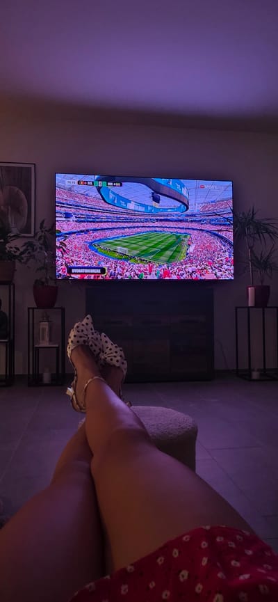 The image shows a woman's legs crossed, resting on a footstool, as she watches a soccer game on a large television screen. The screen displays a stadium filled with spectators and a soccer match in progress, with a score overlay indicating "BEL 0" and "IRI 0" at 70:10. A banner across the screen reads "HYDRATION BREAK". The room is dimly lit, with a purple hue cast from overhead lighting. In the background, there are two tall, black metal shelving units with decorative items and plants. One shelf holds a framed picture. The floor appears to be tiled. The overall setting suggests a relaxed evening at home, possibly in Lieli, Switzerland, given the user's prompt.