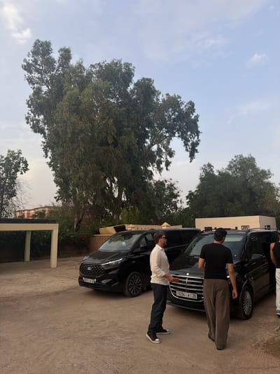 In Al Ouidane, Morocco, a scene unfolds outdoors with two black vans parked on a dirt lot. The van on the left, a Ford Transit, is facing slightly right, its front visible, displaying a license plate reading "60751 | J | 26". A man wearing a white long-sleeved shirt and dark jeans stands to its left, holding an orange object. The van on the right is a Mercedes-Benz, also facing slightly right. A man in a black t-shirt and loose-fitting grey pants walks away from it, towards the right side of the frame. Another person, partially visible, is near the open rear door of the Mercedes. Behind the vans, a building with a light-colored facade and dark-trimmed windows is visible. Lush green trees, some quite large and with dense foliage, dominate the background, suggesting a rural or semi-rural setting within or near Al Ouidane. The sky is overcast with light clouds.