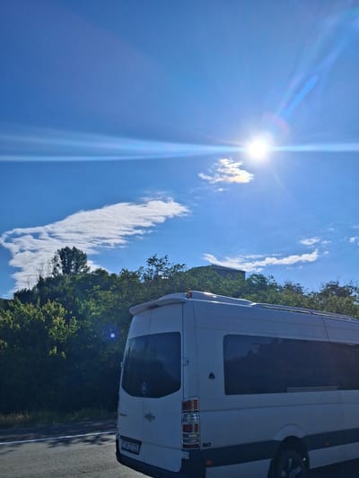 A white Mercedes-Benz Sprinter van is parked on the side of a road in Malishka, Armenia. The van is facing away from the camera, with its rear end visible. It has tinted windows and a license plate that reads "99 ZZ 222". The van is parked in front of a dense green tree line. In the background, a building can be seen peeking through the trees. The sky is bright blue with a few wispy white clouds. The sun is shining brightly, creating lens flare in the image. The overall scene appears to be a sunny day in a rural or semi-rural area.