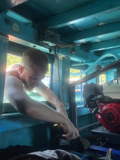The image captures a scene of a man engaged in maintenance or repair work within a blue-painted wooden structure, possibly the interior of a boat or a small utility room. The perspective is looking down into the workspace.
In the lower-center of the frame, a shirtless man with a beard is visible, leaning forward and looking intently at something out of view below him. His arms are extended, and his right hand appears to be manipulating a tool with a yellow handle, resembling a screwdriver, near a black bag.
To the upper right of the man, a red and silver engine, clearly branded "HONDA," is mounted on a metal frame. A light blue, corrugated flexible hose or tube arches across the upper-center of the image, connecting to or running near the engine.
On the left side of the frame, attached to a blue wooden beam, an electrical component resembling a circuit breaker panel or switch box is installed, with wires connected to it and a flexible conduit leading away. The wooden structure itself is composed of multiple blue-painted planks and beams, showing some signs of wear and tear.
Notable text visible includes "HONDA" on the engine and "S.W.A.T." in white letters on the black bag near the man's right hand. Bright sunlight streams into the space, creating strong highlights on the man's skin and some of the surfaces, suggesting it is daytime and an opening is letting light in. A small part of another person's arm or shoulder is visible on the far right edge of the image. The overall impression is one of focused manual labor in a functional, possibly industrial or marine, environment.