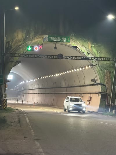 A white SUV drives into Tunnel No. 5, identified by a green sign that also indicates the tunnel's length as 740 meters. A traffic signal above the tunnel entrance shows a green downward arrow, a red circle with the number 60, and a red X, suggesting speed restrictions and an open lane. The tunnel interior is illuminated by a row of lights along its arched ceiling and the headlights of the approaching vehicle. The tunnel walls are covered in a textured material, possibly concrete or rock, with some areas showing moss or vegetation. The roadway is paved and marked with a white line. The scene appears to be at night or in low-light conditions, with the surrounding environment outside the tunnel obscured by darkness and possibly fog.