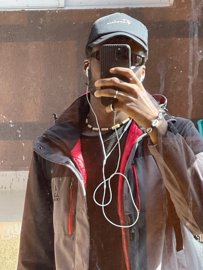 The image is a selfie taken by a young Black man in what appears to be an urban outdoor setting. He is wearing a black baseball cap, sunglasses, a black t-shirt, and a grey and black jacket with red accents. He has white earbuds in and is holding up his smartphone to take the picture. He is also wearing a beaded necklace and a bracelet made of shells. The background is a textured brick wall. The lighting suggests it is daytime. There is no indication of the specific location being Rahwol Chom, Nigeria.