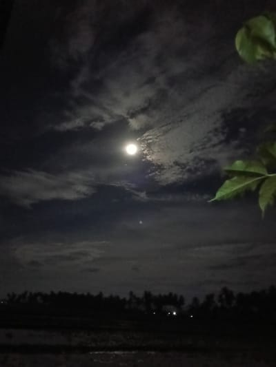 This is a photograph taken at night in Blangkutacoh, Indonesia. The dominant subject is a bright full moon illuminating a cloudy sky. The clouds are wispy and appear to be moving, creating a dynamic texture in the atmosphere. In the foreground, a silhouette of trees and foliage is visible against the night sky. The bottom portion of the image shows a dark, indistinct landscape, likely fields or ground, with some reflective surfaces that could be water. A few faint lights are visible in the distance, suggesting a settlement or scattered dwellings. The overall mood is serene and somewhat mysterious, typical of a moonlit night. The image captures a natural landscape scene under the moonlight. There is no visible text in the image.