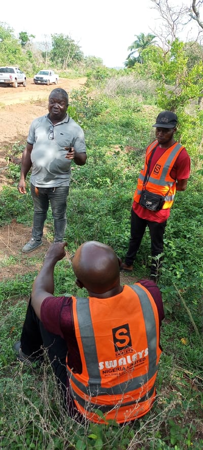 Three men are seen outdoors in a natural, possibly undeveloped, environment in Otunade, Nigeria, during daytime. The scene is dominated by lush green vegetation, including bushes, trees, and tall grasses, with a dirt track visible in the background where two white pickup trucks are parked.

In the foreground, a bald man with dark skin is seated on the ground, facing away from the camera. He wears a maroon long-sleeved shirt and an orange safety vest. On the back of his vest, a black logo with an 'S' and the text "SWALEYS NIGERIA LIMITED BUILDING & ENGINEERING CONTRACTOR" is visible. He is pointing forward with his right hand, seemingly directing attention to something.

Standing on the left, an older man with a sturdy build wears a light grey polo shirt, dark jeans, and grey sneakers. He has glasses hooked onto his shirt collar and holds a dark-colored phone in his right hand, gesturing as if speaking or explaining something.

To the right, another man stands, wearing a red t-shirt, black pants, and a black baseball cap with "POLO" written on it. He also wears an orange safety vest and a black shoulder bag across his body. He is looking down and slightly to his left, seemingly engaged in the discussion or observing what the seated man is pointing at.

The men appear to be involved in a professional discussion or survey of the area, possibly related to construction or development, given the safety vests and the visible company name on one of them. The lighting suggests it is bright daylight, possibly mid-morning or late afternoon.