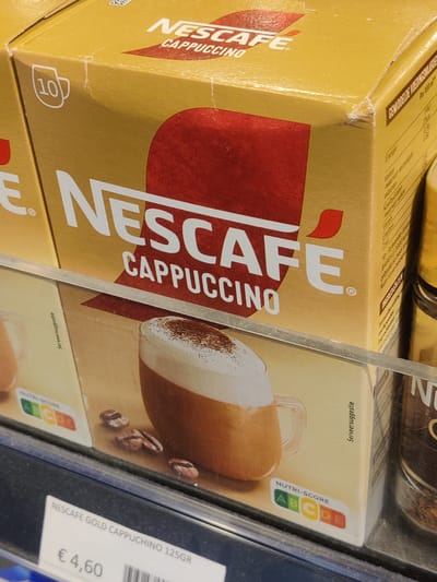The image shows a close-up of a shelf display in a retail setting, likely a supermarket, in Eijsden, Netherlands. The primary focus is on a box of Nescafé Gold Cappuccino, 125g, priced at €4.60. The Nescafé box is predominantly yellow with red accents and features the Nescafé logo, the word "CAPPUCCINO," and a depiction of a frothy cappuccino. A "10" in a circle indicates the number of servings. A Nutri-Score badge is visible on the packaging, showing a grade of 'C'.
To the left of the Nescafé box, another partial box of Nescafé is visible, also with the "NESCAFÉ" branding. To the right, a glass jar with a black lid, likely containing instant coffee or a similar product, is present. The products are displayed on a clear acrylic shelf, with a dark blue shelf edge or frame visible at the bottom. The lighting suggests an indoor retail environment, with overhead lighting casting minimal shadows. The overall scene is static, with no people or actions depicted. The background is slightly blurred, hinting at other products on shelves behind the main subject.