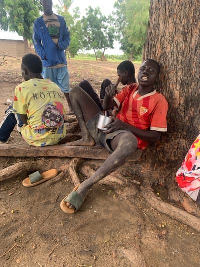 Four individuals, appearing to be young men or boys, are seated on the ground in a shaded area under a large tree. The ground is dirt and covered with exposed tree roots. In the foreground, one individual is wearing a yellow t-shirt with a graphic print and text that reads "BÔTT N" followed by illegible text. Another individual to his left is wearing a blue jacket over a tan and white striped shirt, and light blue shorts. To the right, a third individual is wearing a red and white striped shirt and dark shorts, with a metal cup in his hand. He is looking towards the camera with a wide smile, showing his teeth. The fourth individual is partially visible, sitting behind the one in the red shirt.

The scene appears to be outdoors, likely in a rural setting given the dirt ground and sparse vegetation in the background, which includes trees and what looks like a field. The lighting suggests it is daytime, possibly overcast or late afternoon, as the light is diffused. The people are dressed casually, with one pair of green and brown sandals visible on the ground near the tree roots. A piece of fabric with a red, white, and blue pattern is also visible at the top right of the image. The overall impression is one of a relaxed gathering.