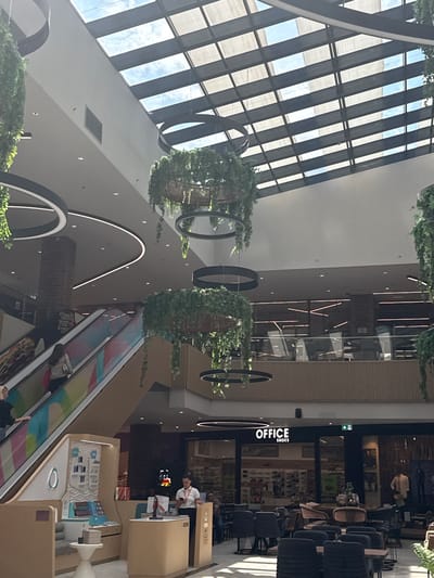 The image captures the interior of the Grand Centar shopping mall in Ilidža, Bosnia and Herzegovina. The scene is brightly lit by natural light filtering through a large, multi-paneled skylight. Suspended hanging plants with circular black frames are a prominent decorative element, adding a touch of greenery to the modern architecture.
In the foreground, a modern retail kiosk is visible, featuring displays of small electronic devices and branded signage. A woman in a white shirt and black skirt stands behind the counter, looking at her phone. To the left, a colorful escalator ascends, with a woman in a light-colored jacket carrying a red bag walking up.
Further into the mall, a restaurant or cafe area is set up with tables and chairs. In the background, a store with the prominent sign "OFFICE SHOES" is visible. The overall environment suggests a busy commercial space with a contemporary design, blending retail, dining, and architectural elements.