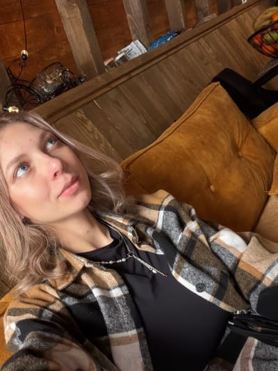 An individual is depicted reclining indoors on a mustard yellow upholstered sofa in Доња Брезна, Montenegro. The subject is a young adult female with light-colored hair and blue eyes, looking upwards and to the viewer's left. She wears a black top beneath an open plaid shirt jacket with beige, brown, black, and white patterns. Two layered necklaces are visible: one with small white beads and another silver-toned chain. The background features exposed wooden ceiling beams and paneled walls. On an elevated shelf in the upper left, a decorative metal bicycle-shaped object with a basket and two small hanging light bulbs are visible. Further back, various packaged items, including a blue bag, are stacked on a shelf. In the upper right, a fruit bowl containing assorted fruit is partially visible.