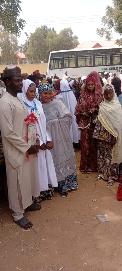 The image depicts an outdoor gathering in Sani Mai Nagge, Nigeria. In the foreground, from left to right, stand a man and four females. The man wears a light beige long tunic and matching cap, and dark sandals. He holds the hand of the woman next to him, who is dressed in a white head covering and long white garment. She wears a blue and white medallion around her neck and holds a framed certificate adorned with red ribbons. Henna is visible on her hands. Next to her, a woman in a blue and white patterned head covering and a grey embroidered top over a blue patterned skirt stands smiling, wearing dark sandals. To her right, a woman in a red head covering and a long red floral dress holds a string of beads in her left hand, wearing silver sandals. A younger female stands furthest right in the foreground, wearing a beige head covering with gold embroidery and a long beige and gold patterned dress, also with silver sandals.
The ground is light brown dirt with scattered small twigs and paper debris. In the midground, a crowd of numerous individuals, predominantly adults in traditional attire and head coverings, extends behind the main subjects. In the background, a large white bus is visible, with "GIRL CHILD IN" printed on its side. Behind the bus, a fence topped with barbed wire runs horizontally. Further back, trees and a building with a reddish roof are partially visible under a bright sky. The scene appears to be a formal event or celebration.