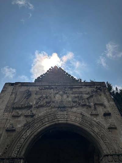 The image shows a low-angle view of the stone facade of the San Andrés Calpan church in Mexico, looking up towards a bright sky with scattered clouds. The facade features intricate stone carvings, including religious figures and decorative motifs. A triangular pediment crowns the structure, also adorned with detailed stonework. The sun is visible behind some of the clouds, casting a bright light and creating a silhouette effect on the top of the facade. Trees are visible on the right side of the image, framing the building. The overall impression is one of ancient architecture set against a natural backdrop.
