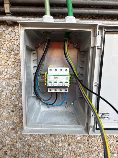 An outdoor electrical junction box is shown, open and mounted on a pebbled wall. Inside, a triple-pole circuit breaker labeled "Schneider Electric 30F3" is visible. Multiple colored electrical wires, including brown, blue, yellow-green, and black, are connected to the breaker and run through the box. Two green conduits enter the box from the top, one on each side, and the wires emerge from them. The inside of the box appears to have some dust or debris. The exterior of the box is gray plastic. The background consists of a wall made of small, multi-colored pebbles and some metallic conduit pipes. The scene suggests an electrical installation or maintenance activity. The location is the municipality of Zwijndrecht, Belgium.