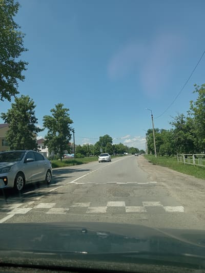 A street scene in Otradnaya, Russia, under a clear blue sky. The asphalt road features a pedestrian crossing marked with white squares. On the left side of the road, a silver sedan is parked, its front wheel near the crossing. Further down the road, other vehicles are visible in the distance. Trees line both sides of the road, and buildings can be seen behind the trees on the left. A metal fence runs along the right side of the road. The overall scene depicts a typical day in a Russian town.