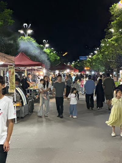 The image depicts a bustling night market scene in Eshan Yi Autonomous County, China. A diverse crowd of people, including families with children, stroll along a paved pathway lined with food stalls and trees adorned with decorative lights. Smoke billows from one of the stalls, suggesting cooking is in progress. Several individuals are actively engaged in shopping or dining. The lighting suggests it is nighttime, with streetlights and stall illumination creating a vibrant atmosphere.