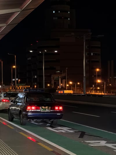 A nighttime street scene features a dark blue "Yaesu Taxi" with illuminated rear lights, parked near a green lane with a white road marking. Japanese text on the taxi reads "3/1 (Sun) Tokyo Marathon, entrance closed." Buildings line the dark background with streetlights visible along the road. A license plate is partially readable with "500" and "82" visible. Other cars are parked ahead of the taxi.