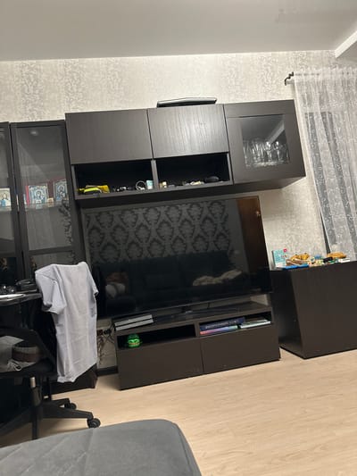 The image depicts an indoor living room scene. A large dark brown entertainment center dominates the right side of the frame, featuring several cabinets and shelves. A large flat-screen television is mounted on the wall, displaying a dark, patterned background. To the left of the entertainment center, a black office chair with a light gray t-shirt draped over its back is visible. A portion of a gray sofa is visible in the foreground. On a shelf of the entertainment center, a small, green, cartoon-like object with wide eyes and a grinning mouth is placed. The walls are covered with textured wallpaper. To the right, white sheer curtains are drawn. The floor is covered with light wood-look laminate flooring. The room appears to be situated in a residential apartment in the city of Kinel, Russia.