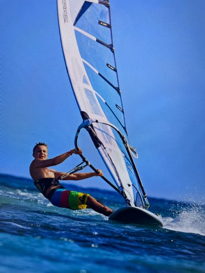 A man is windsurfing on blue water under a clear blue sky. He is gripping the boom with both hands, his body suspended beneath it. He is wearing colorful board shorts. The windsurf board is slicing through the water, creating spray. The sail is blue and white. The photo appears to be taken at an angle. It is not possible to determine the exact location within the city of Bacong, Philippines from this image alone.