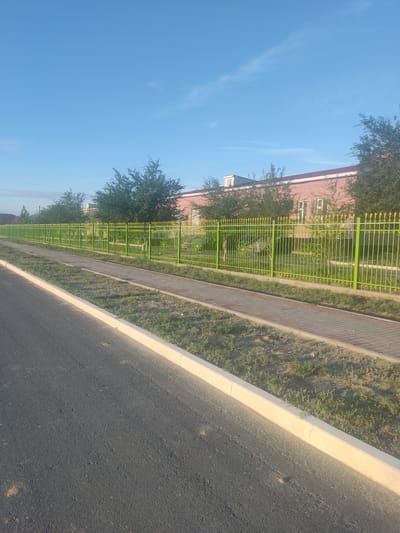 The image depicts a scene in the city of Aral, Kazakhstan. In the foreground, a dark asphalt road with a white concrete curb runs diagonally from the bottom left corner towards the top right. Adjacent to the road is a pedestrian walkway made of light-colored paving stones, bordered by a strip of dry, sparse grass.
Beyond the walkway is a bright green metal fence with ornate gold finials. The fence encloses an area with more grass and some young trees. In the background, a long, single-story building with a reddish-brown brick facade and a white roofline is visible. Several trees with dense green foliage are situated in front of the building, partially obscuring it. The sky above is a clear, bright blue with a few wispy white clouds. The overall scene suggests a well-maintained public area, possibly in a residential or educational district of Aral. There are no people or animals visible in the image.